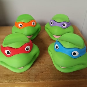 May include: Four green turtle heads with different colored eye masks: orange, purple, red, and blue. Each has white eyes and a smiling pink mouth. The heads are arranged on a wooden surface.