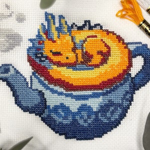 Peut inclure: Une théière bleue et blanche avec un dragon orange et jaune enroulé qui dort à l'intérieur. Le dragon a une crinière rouge et orange.