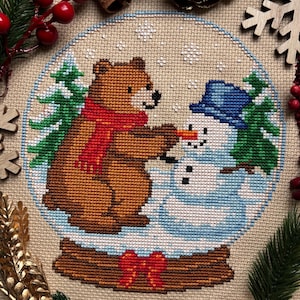 Può includere: Design a punto croce di un orso con una sciarpa rossa e un pupazzo di neve all'interno di una palla di neve. L'orso tiene il naso di carota del pupazzo di neve. La palla di neve poggia su una base di legno con un fiocco rosso.