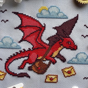 Puede incluir: Diseño de punto de cruz con un dragón rojo que lleva una bolsa marrón, volando junto a nubes azules y sobres amarillos. El dragón tiene grandes alas rojas sobre un fondo azul claro. Pequeños pájaros negros están dispersos.