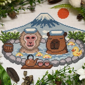 Può includere: Design a punto croce con una scimmia e un orso che si rilassano in un onsen, o sorgente termale, con il Monte Fuji sullo sfondo. La scena comprende una teiera, tazze e una paperella di gomma. La tavolozza dei colori comprende blu, marroni e arancioni.