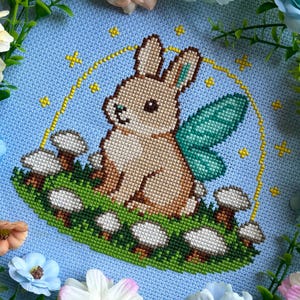 Può includere: Un disegno a punto croce con un coniglietto marrone con ali di fata turchesi, seduto tra funghi su una macchia verde. Il coniglietto è all'interno di un cerchio giallo con stelle gialle su sfondo azzurro.