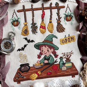 Può includere: Design a punto croce con una strega con cappello e vestito verdi, che lavora a un tavolo con un calderone, scope e la parola "OPEN". Pipistrelli e piante sospese si aggiungono al tema di Halloween. Un orologio da tasca è nell'angolo.