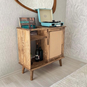 May include: A wooden record player stand with a light blue record player on top. The stand has a woven rattan door and a shelf for storing records and bottles. The stand has a mid-century modern design.