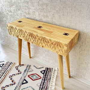 Könnte beinhalten: Ein heller Holz-Konsolentisch mit aufwendigen geometrischen Schnitzereien. Der Tisch hat eine rechteckige Platte und vier Beine. Der Tisch steht auf einem gemusterten Teppich.