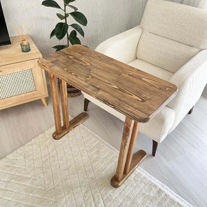 May include: A wooden side table with a rectangular top and angled legs. The table is made of light brown wood with visible grain. The legs have a unique design, and the table is placed on a cream-coloured rug.