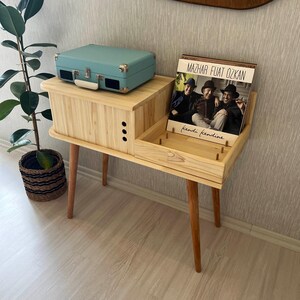 May include: A light blue record player sits on a wooden record stand with a record album titled "Mazhar Fuat Ozkan kendi kendine" on the right side of the stand.