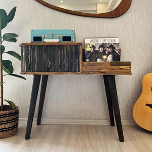 May include: A wooden record player stand with a black finish and black legs. The stand has a shelf for records and a drawer for a record player. A turquoise record player is sitting on the top shelf. A record album with the text "Mazhar Fuat Ozkan" is on the lower shelf.