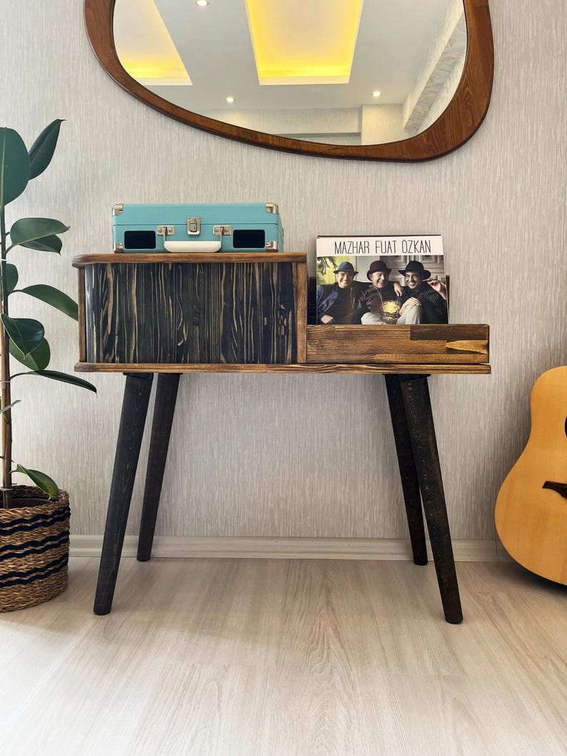 May include: A wooden record player stand with a black finish and black legs. The stand has a shelf for records and a drawer for a record player. A turquoise record player is sitting on the top shelf. A record album with the text "Mazhar Fuat Ozkan" is on the lower shelf.