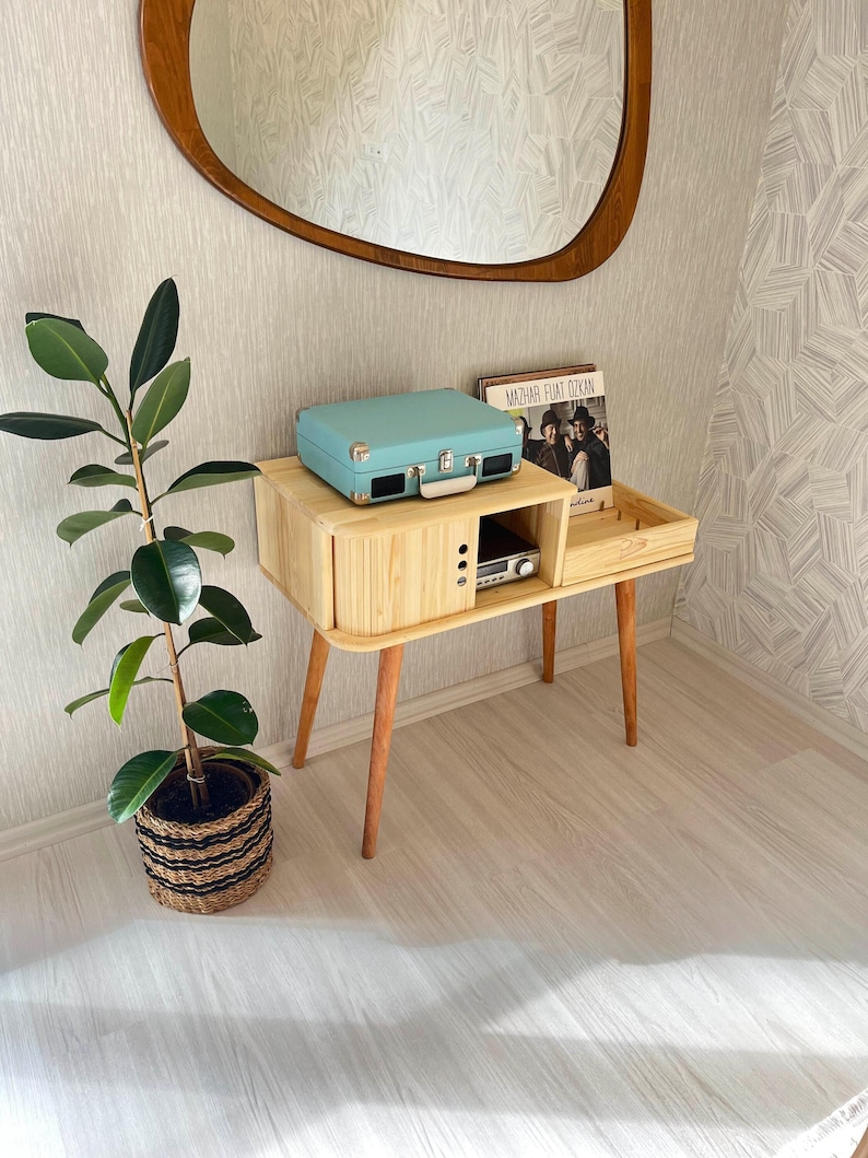 May include: A light wood record player stand with a light blue record player on top and a record album on the shelf. The stand has four legs and a small drawer. A potted plant is on the floor next to the stand.