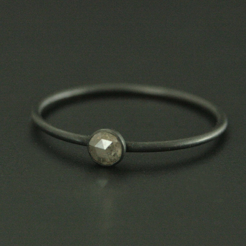 May include: A simple black band ring with a single, round, gray gemstone set in the center.
