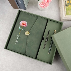 May include: A green gift box set containing a journal, pen, and envelope closure. The journal features a floral design with a pink carnation and the name "Garrett" inscribed on the front.
