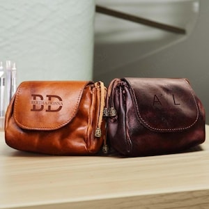 May include: Two leather pouches in shades of brown and burgundy sit side-by-side. The lighter pouch has the initials "DD" and the text "BERTHA.DONNA" printed on the front. The darker pouch has the initials "A L" printed on the front.