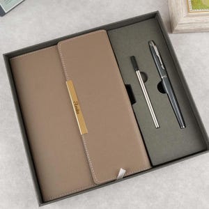 May include: A brown leather-bound journal with a gold nameplate that reads "Julia" and a black pen and a silver pen in a black box.