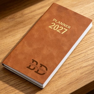 May include: A rectangular tan planner with a smooth finish, featuring the gold-embossed text PLANNER 2027 at the top and a dark brown monogram logo with the name BERTHA.DONNA printed at the bottom, with white pages visible along the side edge.