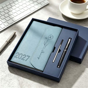 May include: A light blue 2027 planner with a floral line drawing and the name Jenny embossed on the cover, presented in a dark blue gift box alongside a black ballpoint pen and a metal ink refill, all arranged in a structured display set.