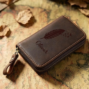 May include: A brown leather wallet with a zip closure. The wallet has a feather design and the name "Oliver" engraved on the front. The rectangular wallet appears to be made of high-quality leather. The zip pull is also leather.