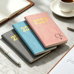 May include: Three rectangular diaries stacked diagonally, featuring a pink, blue, and grey cover. Each diary is embossed with the year 2027 in gold foil. The top pink diary is personalised with the letters CK and a small heart symbol in black.