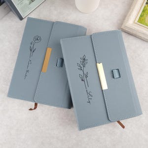 May include: Two gray leather-bound journals with gold accents and floral designs. The journal on the left has a rose design and the text "Rose Bud". The journal on the right has a lily design and the text "Lily".