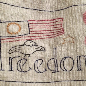 May include: A white fabric with a blue border and embroidered design. The design includes an American flag, a bird, and the word "freedom".