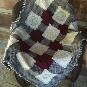 May include: A handmade quilt with a blue and white gingham pattern and white squares with embroidered designs. The quilt is folded and draped over a wicker chair.