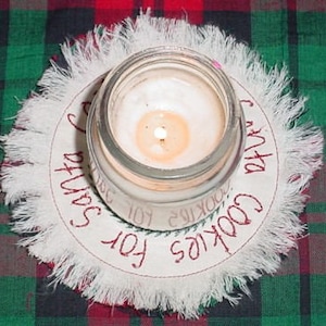 May include: White fabric circle with frayed edges and red embroidery that reads "Cookies for Santa". A clear glass jar with a lit candle sits in the center.