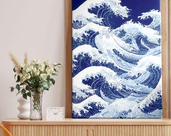 Op de afbeelding: Een ingelijste canvas print met een gestileerde afbeelding van oceaangolven in blauw en wit. Het kunstwerk wordt getoond op een houten kast, met een moderne stoel en decoratieve items op de achtergrond.
