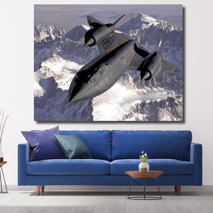 May include: A large canvas print of a black SR-71 Blackbird aircraft soaring over snow-capped mountains. The high-altitude reconnaissance aircraft is angled slightly downward, with its distinctive shape and red accents visible. The artwork is displayed above a blue sofa.