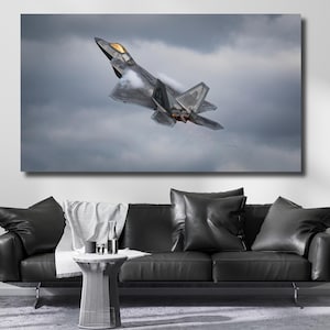 Op de afbeelding: Een canvas print van een grijze F-22 straaljager die door een bewolkte lucht vliegt. De straaljager staat omhoog, met zichtbare details van zijn vleugels en staart. Het kunstwerk staat boven een zwarte leren bank.