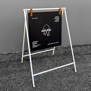 Può includere: Un cavalletto pubblicitario bianco a forma di A con una lavagna rettangolare nera sospesa da due cinghie in pelle marrone. La lavagna presenta testo bianco, tra cui "NOCT. COFFEE", "NOW OPEN" e dettagli aziendali con un'illustrazione.