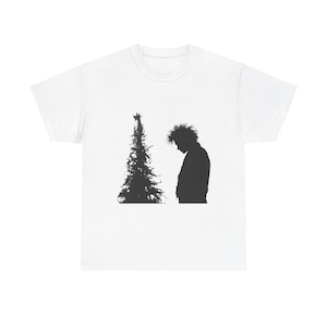 May include: White t-shirt featuring a black silhouette of a Christmas tree and a person. The person is standing next to the tree, with their head bowed. The design is simple and graphic.