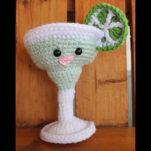 May include: A crocheted green margarita glass with a white rim and a white stem. The glass has a cute face with black eyes and a pink mouth. It has a green crocheted lime wedge on the rim.