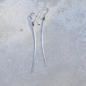 Könnte beinhalten: Ein Paar lange, dünne silberne Ohrringe. Die Ohrringe bestehen aus einem einzigen Stück Metall, das zu einer gebogenen Form gebogen ist.