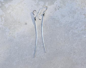 Può includere: Un paio di orecchini lunghi e sottili in argento. Gli orecchini sono realizzati in un unico pezzo di metallo piegato in una forma curva.