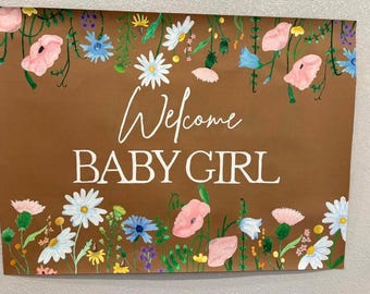 May include: Brown sign with the words "Welcome BABY GIRL" in white script. The sign is decorated with watercolor flowers in pink, blue, white, and yellow. The flowers are arranged around the edges of the sign.