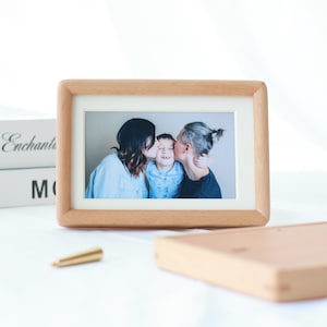 Könnte beinhalten: Ein rechteckiger Bilderrahmen aus hellem Holz. Er enthält ein Foto von drei Personen. Ein kleines, goldfarbenes Objekt und ein Holzobjekt befinden sich im Vordergrund.