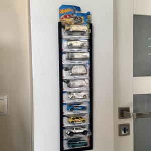 Puede incluir: Un expositor negro montado en una pared blanca, que muestra una colección de coches de juguete Hot Wheels. El expositor contiene varios vehículos en miniatura empaquetados en varios colores, incluyendo amarillo, azul y blanco. La pantalla está cerca de una puerta blanca con un tirador plateado.