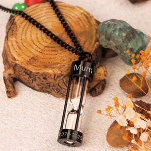 May include: Black hourglass pendant necklace with the word "Mum" and the years "1970-2025" engraved. The pendant is suspended from a black chain. The background includes a wooden slice, rocks, and dried flowers.