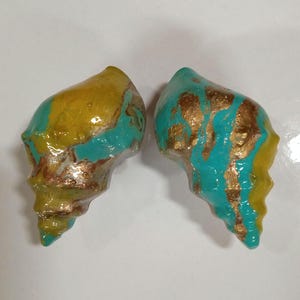 Può includere: Due conchiglie dipinte in tonalità turchese, giallo e oro. Le conchiglie hanno una finitura lucida.
