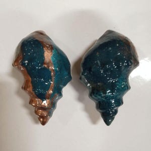 May include: Two ceramic seashell sculptures. The shells are teal with bronze accents. The sculptures have a glossy finish and are displayed against a white background. The shells are a decorative item.