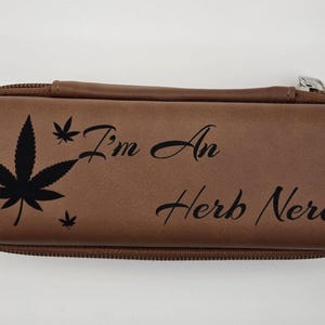 May include: A brown glasses case with a zip. It has a black cannabis leaf design and the words "I'm An Herb Nerd!" printed on it.