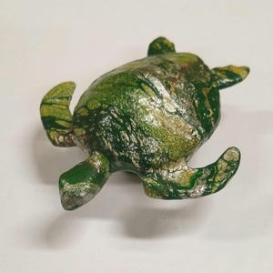 May include: A green and gold painted turtle figurine with a marbled effect. The turtle is lying on its back with its legs and head outstretched.