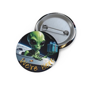 May include: Two round buttons. One button features a green alien holding a blue gift box, emerging from a spaceship. The text "We're Here" is written in yellow. The second button is silver.