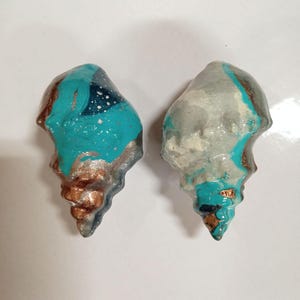 May include: Two seashells painted in a teal, white and gold swirl pattern. The shells are a light teal colour with white and gold accents.