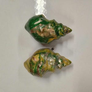 Puede incluir: Dos conchas pintadas con remolinos verdes, dorados y blancos. Las conchas están dispuestas verticalmente, con la concha superior superponiéndose ligeramente a la inferior.