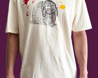 May include: A white t-shirt with a black and white illustration of a rabbit with a red bird and yellow stars on a cream background.