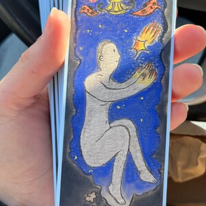Puede incluir: Una ilustración azul y negra de una persona flotando en el aire con una estrella fugaz sobre ella. La persona está extendiendo la mano hacia la estrella. Hay una cama con dos personas en ella debajo de la persona. La ilustración está en una tarjeta blanca con un borde azul.