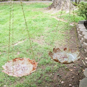 May include: Two hanging ceramic bird feeders suspended by thin metal chains. The left feeder features a raised hexagonal honeycomb pattern with a tan and brown glaze, while the right feeder has a scalloped, petal-like edge with a smooth, glossy finish.