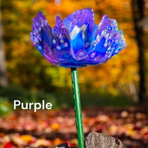 Pu&ograve; includere: Un fiore di vetro viola con accenti blu e bianchi. Il fiore &egrave; su uno stelo verde e si trova su una superficie di legno.