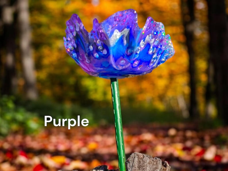 Pu&ograve; includere: Un fiore di vetro viola con accenti blu e bianchi. Il fiore &egrave; su uno stelo verde e si trova su una superficie di legno.